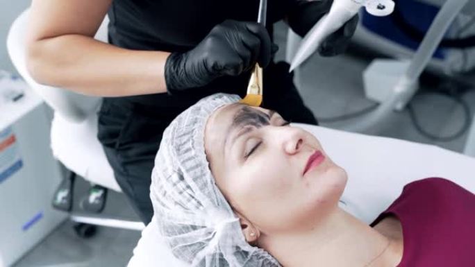 特写美容师在女人脸上的皮肤上涂抹黑碳面膜，慢动作