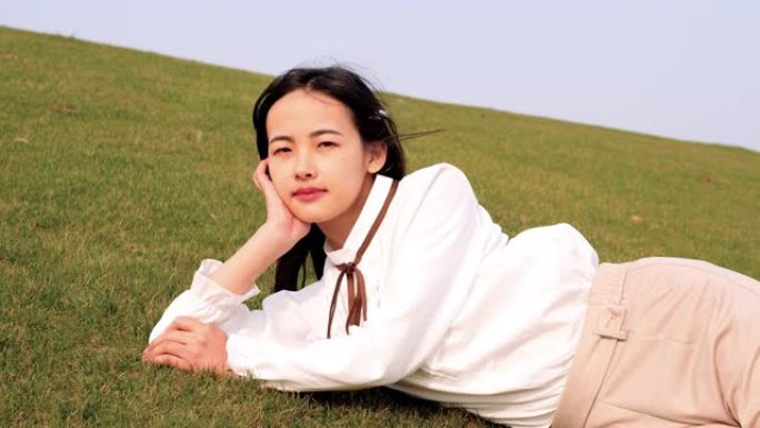 一位美丽的中国年轻女子的肖像躺在绿色的草地上，手握着头，对着镜头微笑，享受春天的微风，4k镜头，慢动