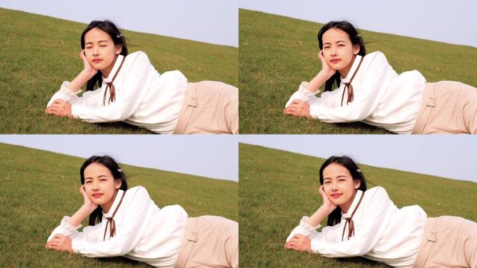 一位美丽的中国年轻女子的肖像躺在绿色的草地上，手握着头，对着镜头微笑，享受春天的微风，4k镜头，慢动