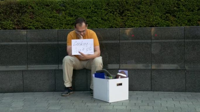 街上有科尔顿的失业者需要工作。被解雇的男子坐在水泥城市的街道上，带着一个装有个人物品的盒子