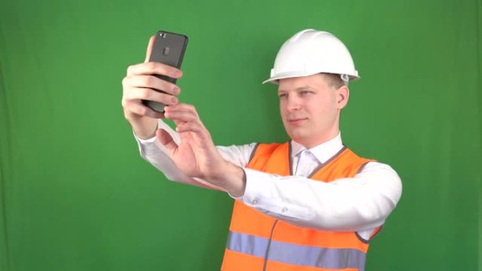 一个穿着信号背心和建筑头盔的年轻高加索男子在建筑工地的电话上自拍，工作场所安全，背景，chromak