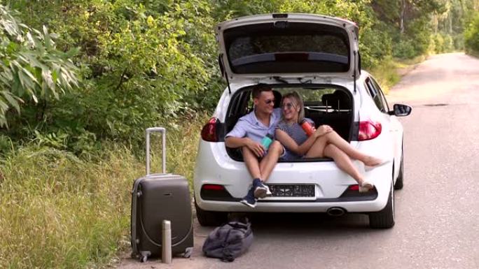 相爱的夫妇坐在汽车后备箱里喝热水瓶里的热茶。