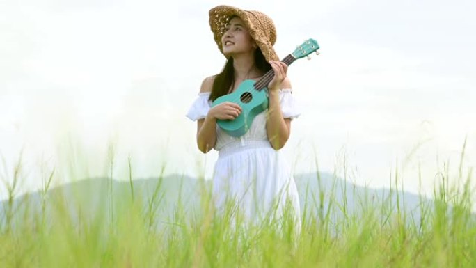 夏天，穿着白色连衣裙的亚洲幸福女人在绿色草地上玩尤克里里吉他，背景是山顶。音乐家美女用乐器放松。乡村