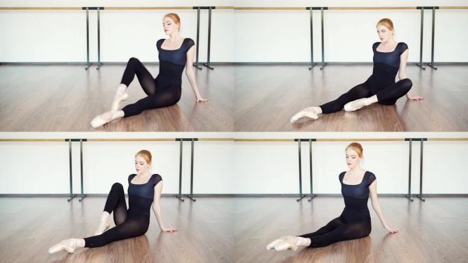 穿着黑色紧身衣和脚尖鞋的优雅年轻芭蕾舞演员的跟踪镜头，坐在芭蕾舞工作室的地板上热身和伸展