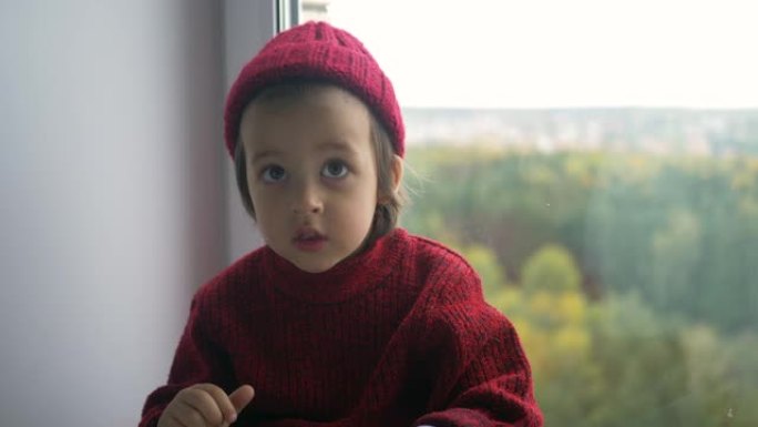 穿着红色毛衣和针织帽的男孩孩子坐在一个大白色窗户上