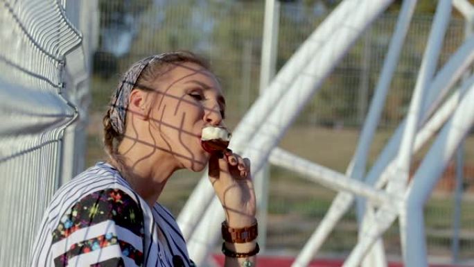 年轻女子靠在栅栏上，手里拿着滑板在篮球场上吃冰淇淋