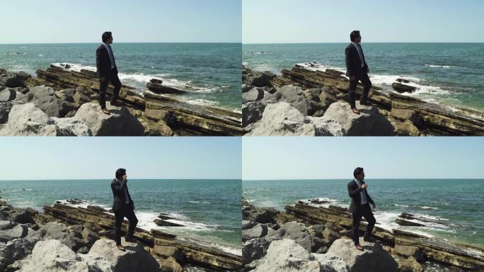 在阳光明媚的夏天，商人站在沙滩上蓝色的岩石上，领带西装和皮鞋，黑色太阳镜。下班后目光思索，坚定而放松