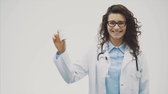 漂亮的年轻女孩医生，穿着医疗礼服。在这段时间里，她处于灰色背景下，手里拿着一瓶白色的药片。在此期间，