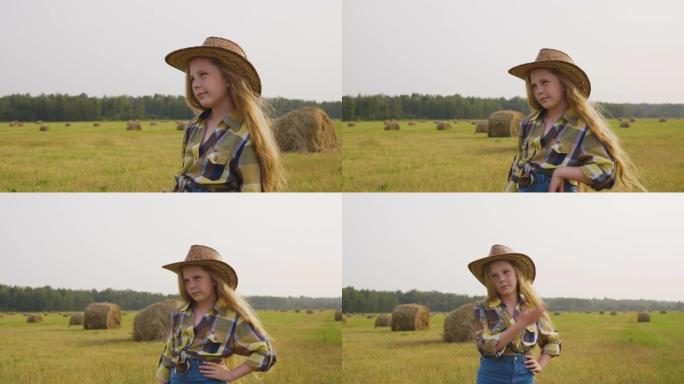 草堆背景下乡村田野上的女牛仔。戴着帽子的年轻奶牛女孩摆前置摄像头。乡村女孩穿着方格衬衫在村子的收割田