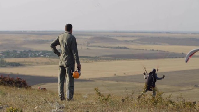 一名身穿工作服的飞行员，手里拿着橙色头盔，站在山坡上，看着语用员起飞。