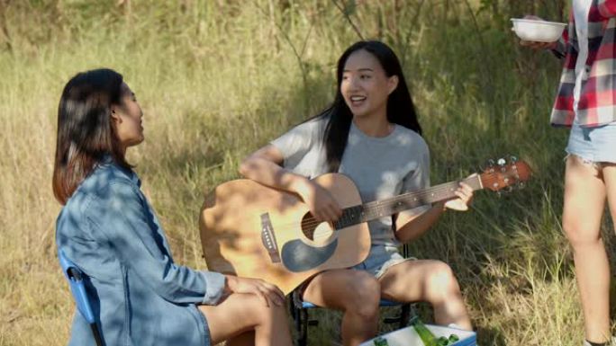 一群年轻的亚洲女性与朋友一起在大自然中露营，一起弹吉他，喝啤酒和碰杯，玩得开心。