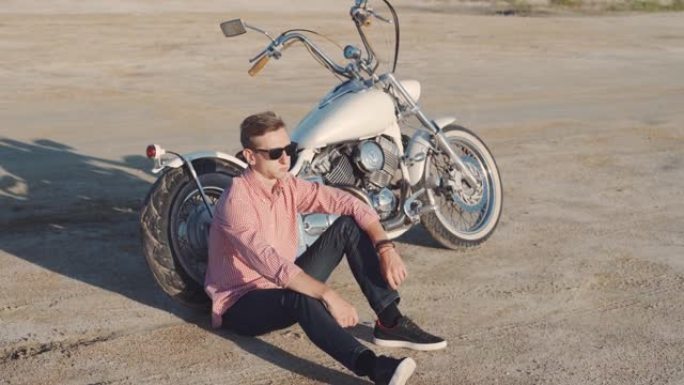 日落时，年轻时尚的摩托车手坐在沙漠路上的摩托车上