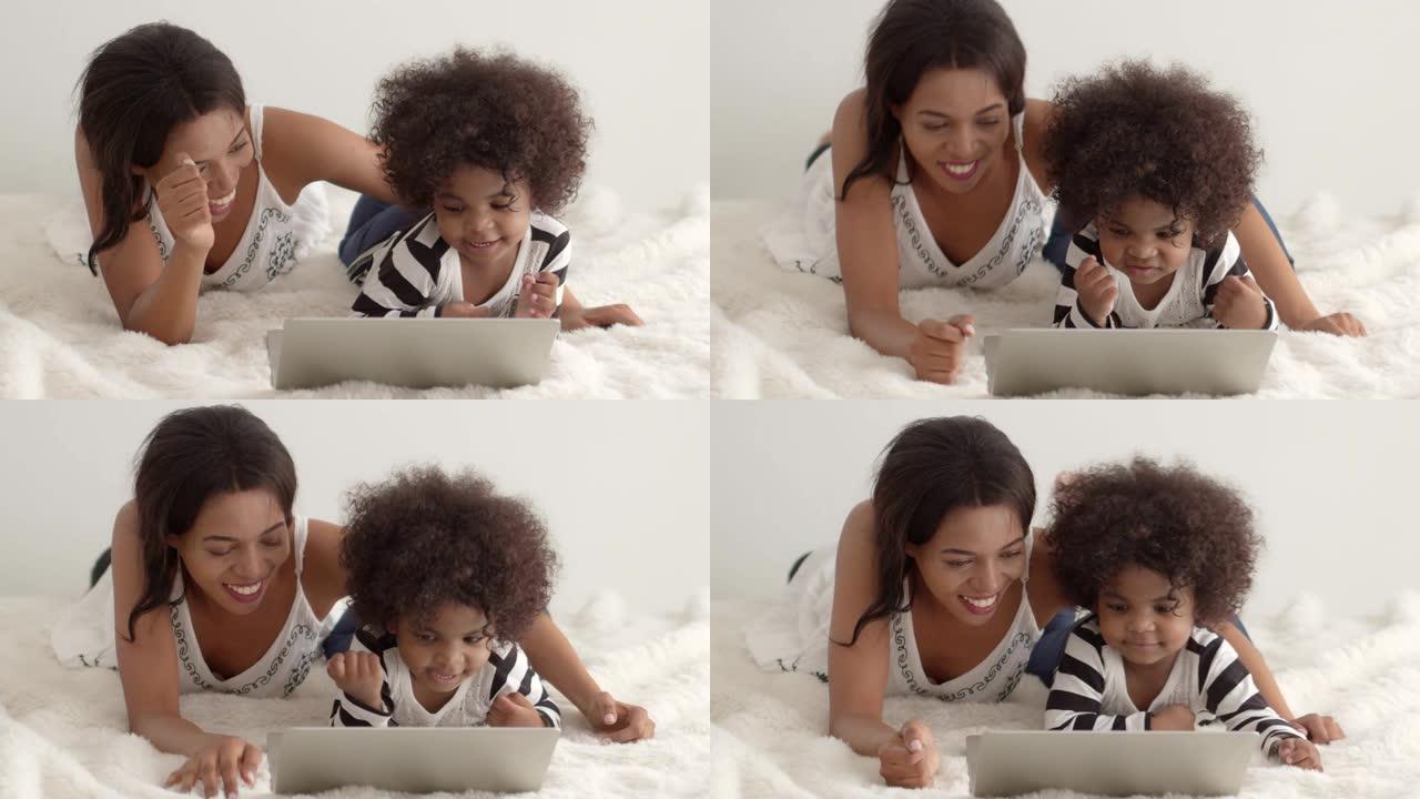 年轻的非洲母亲在笔记本电脑上看卡通或有趣的歌曲，她的儿子在白色床上。慢动作镜头。