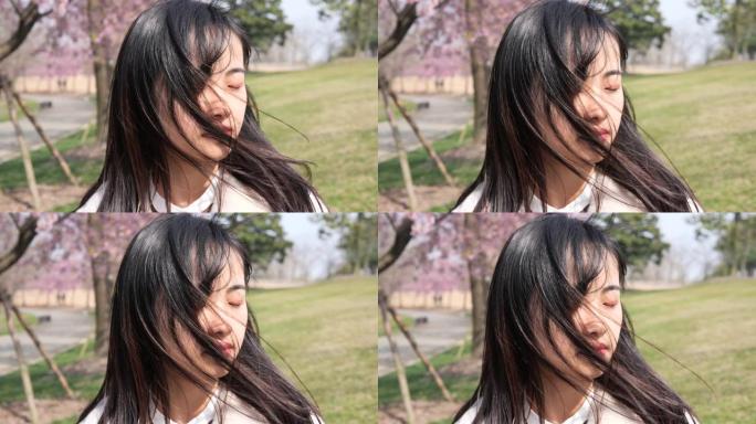 身着粉红色樱花背景的美丽中国年轻女子的头像，闭上眼睛，享受微风吹拂她的黑色长发，春天的美丽，4k，慢
