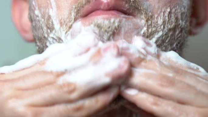 残忍的人呆在镜子前，用剃须放松的洗发水给他没刮胡子的胡子涂肥皂。用电动修剪器准备刮胡子的人。剃光理发