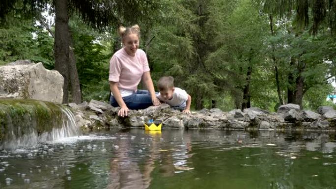 快乐的妈妈和小男孩一起在城市喷泉的水面上吹着黄色的纸船，上面挂着蓝色的旗子。他们微笑和大笑。家庭幸福