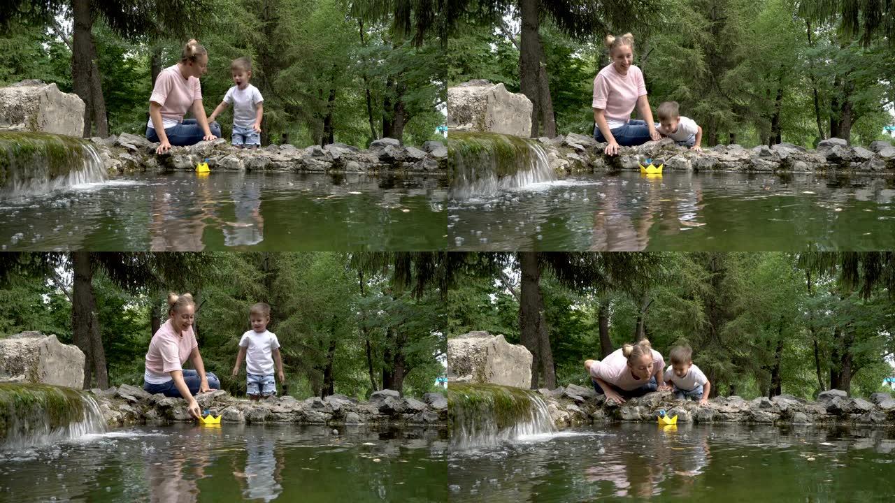 快乐的妈妈和小男孩一起在城市喷泉的水面上吹着黄色的纸船，上面挂着蓝色的旗子。他们微笑和大笑。家庭幸福