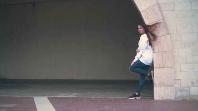 一个留着长发，穿着白色运动衫的女孩，脚踩在建筑物的石拱门上。