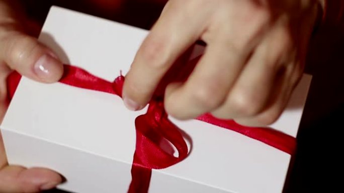 美丽的女孩打开她的礼物。一个女孩美丽的手打开新年礼物，解开红丝带。节日礼物。带红丝带的白色盒子里的礼