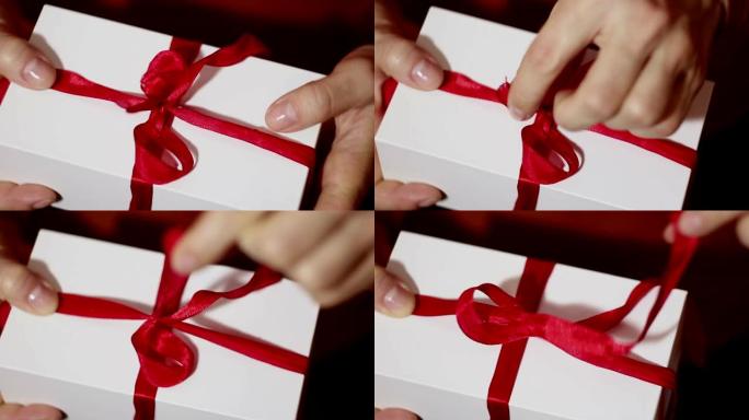 美丽的女孩打开她的礼物。一个女孩美丽的手打开新年礼物，解开红丝带。节日礼物。带红丝带的白色盒子里的礼