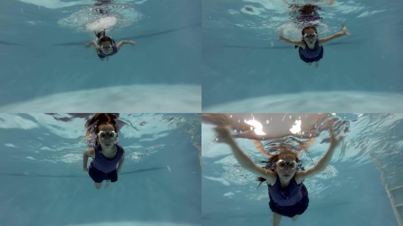 一个快乐的小女孩穿着蓝色连衣裙和游泳眼镜在游泳池水下潜水游泳。她看着镜头微笑。特写。底视图。慢动作。