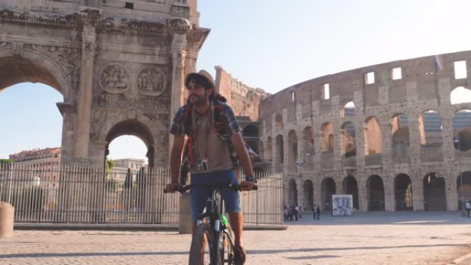 快乐的年轻人游客在日出时在意大利罗马的罗马斗兽场骑着自行车，穿着背包，衬衫和帽子。