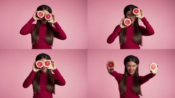 红色年轻美女的肖像，粉色工作室背景上有两半多汁的葡萄柚。健康饮食，节食，抗氧化剂概念