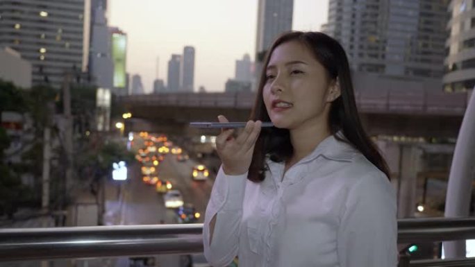在泰国曼谷暮色的现代市中心，快乐的年轻微笑的亚洲女性在智能手机上使用语音识别。通过手机进行概念技术通