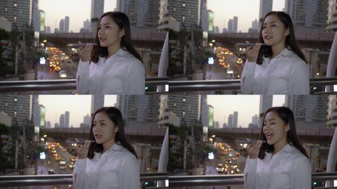 在泰国曼谷暮色的现代市中心，快乐的年轻微笑的亚洲女性在智能手机上使用语音识别。通过手机进行概念技术通