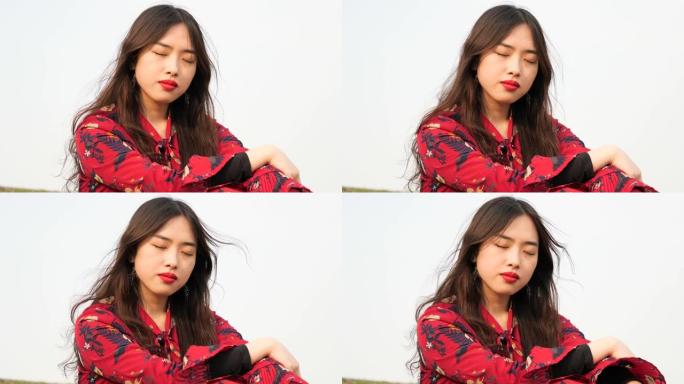 一位穿着红色连衣裙的美丽中国年轻女子的肖像坐在草地上，闭着眼睛，享受微风吹着她的黑色长发，4k镜头，