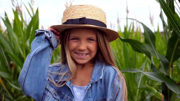 迷人的快乐女孩的肖像看着镜头，在玉米田的背景下拉直她的草帽。留着金色长发的快乐小孩站在草地上大笑。