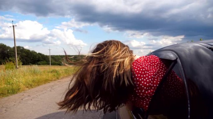 黑发女孩斜靠在车窗外，一边骑车穿过乡间小路一边享受旅行。年轻女子把头伸出移动的汽车，长长的棕色头发随