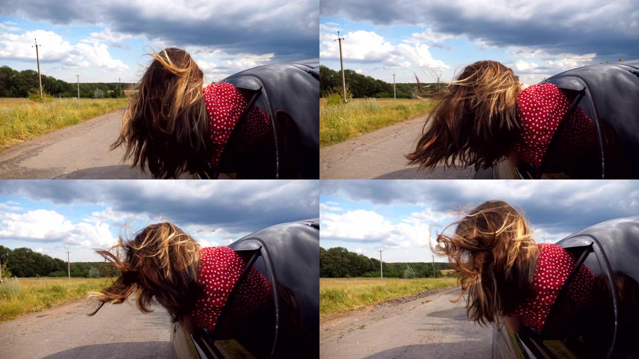 黑发女孩斜靠在车窗外，一边骑车穿过乡间小路一边享受旅行。年轻女子把头伸出移动的汽车，长长的棕色头发随