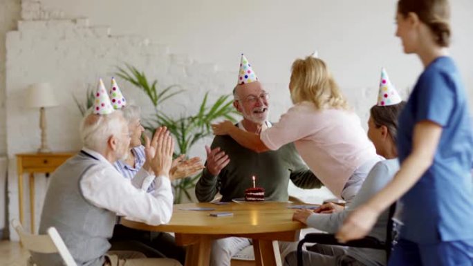年轻护士为老人带来生日蛋糕，与老年朋友在辅助生活家中的派对帽子打牌。快乐的老人收到愿望，拥抱并吹灭蜡