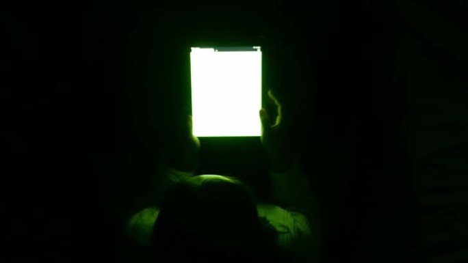 亚洲妇女使用平板电脑，屏幕上的光线在晚上的卧室里照耀着她的脸。