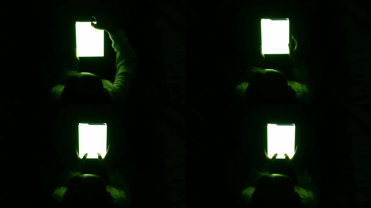 亚洲妇女使用平板电脑，屏幕上的光线在晚上的卧室里照耀着她的脸。