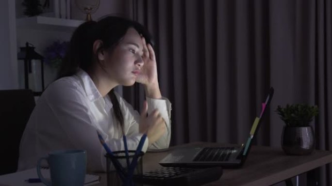 年轻的亚洲单身女学生的坦率工作深夜，在家庭办公室的笔记本电脑或笔记本电脑上解决项目研究问题。亚洲人职