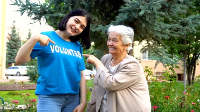 老年女士和年轻志愿者在相机上指着t恤上的志愿者单词