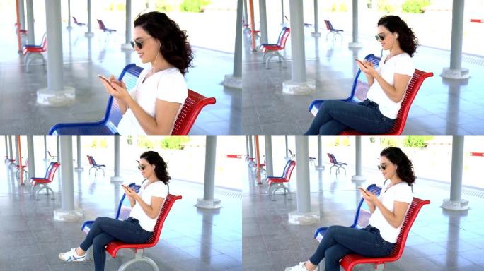 年轻女子在火车站等着用智能手机用耳机听音乐。现代技术，有趣和快乐的人的概念。