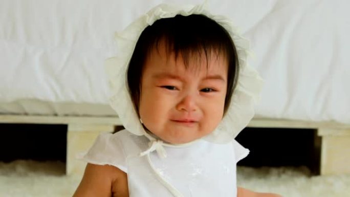 可爱甜美可爱的亚洲宝宝穿着白色连衣裙躺在白色床上哭泣，在舒适的卧室里情绪低落，健康的宝宝概念