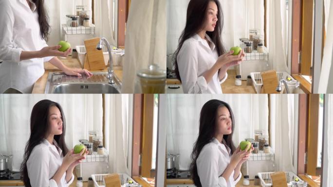 4k中型起重机拍摄孤独的年轻亚洲女子站在厨房里拿着青苹果。不快乐的亚洲少女独自站在厨房窗户旁思考，悲