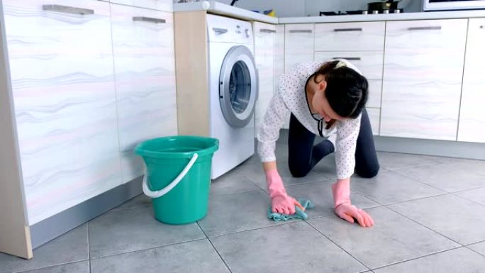 穿着粉红色橡胶手套的疲倦女人用布洗厨房地板。地板上的灰色瓷砖。