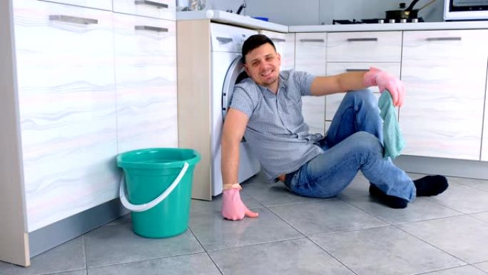 戴着橡胶手套的快乐微笑的男人坐在厨房地板上打扫卫生。