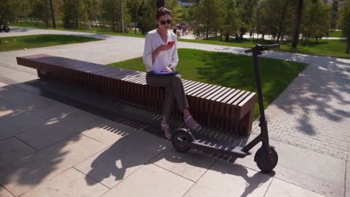 静态拍摄-一个女孩正坐在智能手机旁边，移动生态交通工具。帅气的女性人，阳光灿烂的日子，电动滑板车