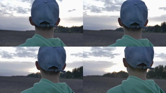 戴着帽子的男孩在傍晚的后视图中看着远处的天空，特写