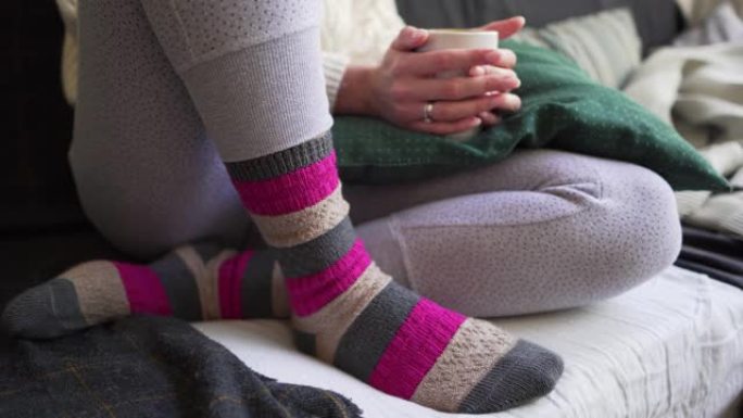 粉红色和棕色的保暖羊毛袜特写-一个女人坐在沙发上，上面放着一杯白杯热饮，上面放着格子和枕头。Lago