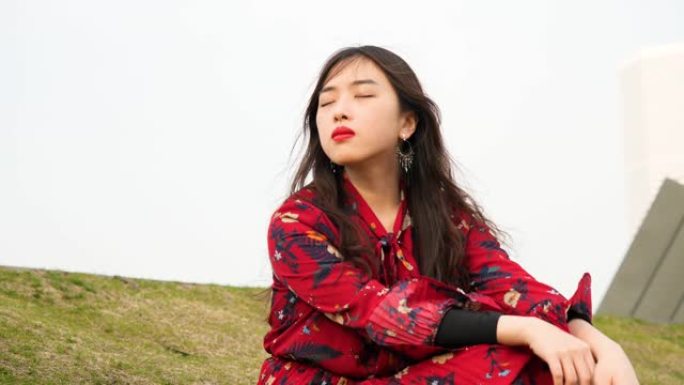一位穿着红色连衣裙的美丽中国年轻女子的肖像坐在草地上，闭着眼睛，享受微风吹着她的黑色长发，4k镜头，