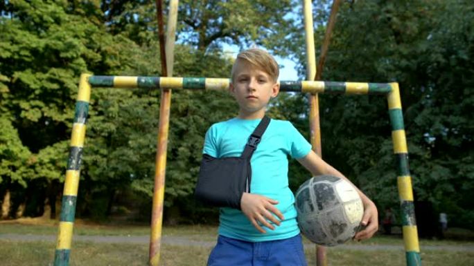 沮丧的男孩穿着肩膀固定的吊索，手臂受伤，拿着足球