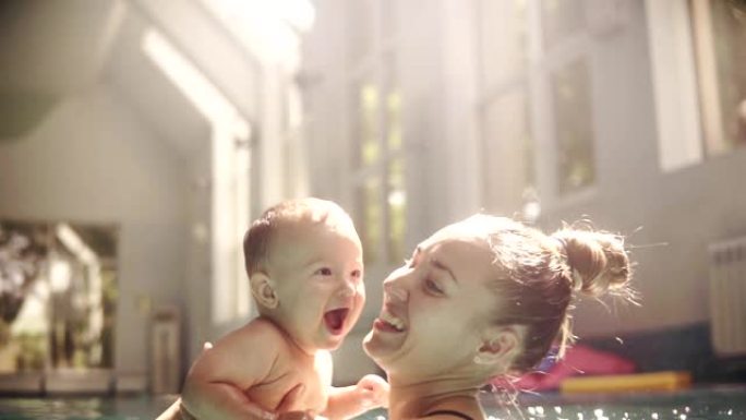快乐的年轻妈妈把婴儿扔进游泳池。孩子的发展。室内游泳池的窗户发出明亮的光线。慢动作