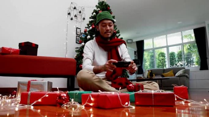 微笑亚洲男子使用手机拍摄照片礼品盒，并上传到互联网在家里的客厅有一棵圣诞树的背景。圣诞节庆祝概念。